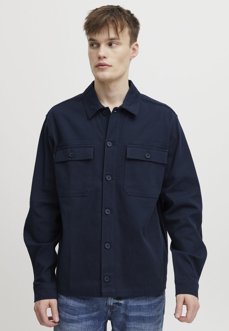 Veste boutonnée bleu marine en coton avec deux poches poitrine, manches longues et col. Tissu texturé au design classique.