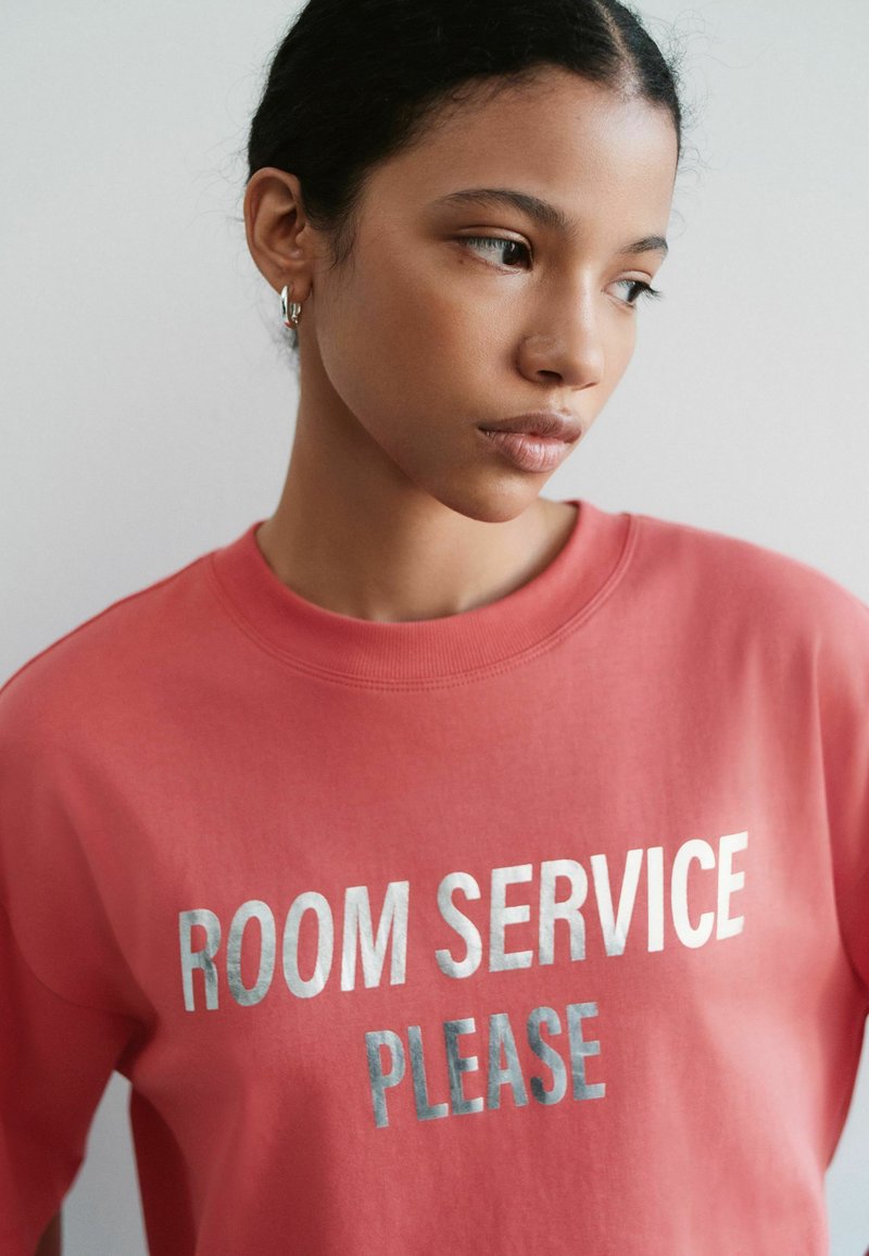 Jeune femme aux cheveux foncés portant un sweat-shirt corail avec un texte argenté indiquant "ROOM SERVICE PLEASE" sur un fond clair uni.