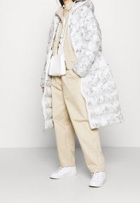 Vadderad grå och vit jacka med huva, tillsammans med en beige outfit som inkluderar lösa byxor och vita sneakers. Innehåller en liten crossbody-väska.