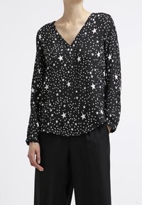 Blouse noire à manches longues et col en V, avec un motif d'étoiles blanches. Tissu léger et texturé avec une coupe décontractée.