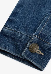 Σκούρο μπλε τζιν ύφασμα με κομμένο τελείωμα, που διαθέτει χρυσή ραφή και μεταλλικό κουμπί με το όνομα της μάρκας σκαλισμένο.