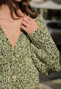 Strikket cardigan i grøn med hvide og sorte detaljer. Har dyb V-hals, bølgede kanter og store træknapper. Blød tekstur.