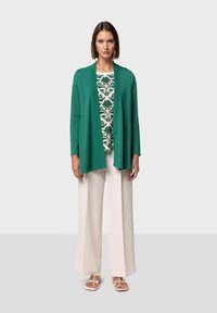 Grön öppen cardigan över en mönstrad vit och grön topp, matchad med krämfärgade vida byxor och lätta sandaler.