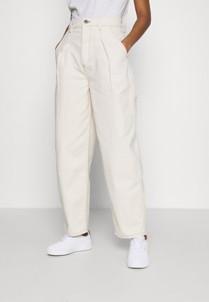 Osoba nosząca szerokie spodnie z wysokim stanem w kolorze kremowym, z kieszeniami, do tego białe trampki i włożona do środka biała koszula.