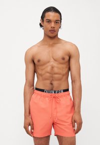 Calvin Klein badeshorts i livlig korallfarge, laget av glatt stoff med justerbar midje og logodetalj i kontrasterende svart.
