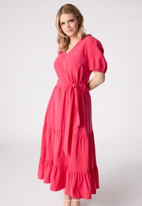 Robe maxi rose à volants avec un décolleté en V, des manches bouffantes et une ceinture nouée. Tissu doux avec un ourlet à volants qui tombe fluidement.