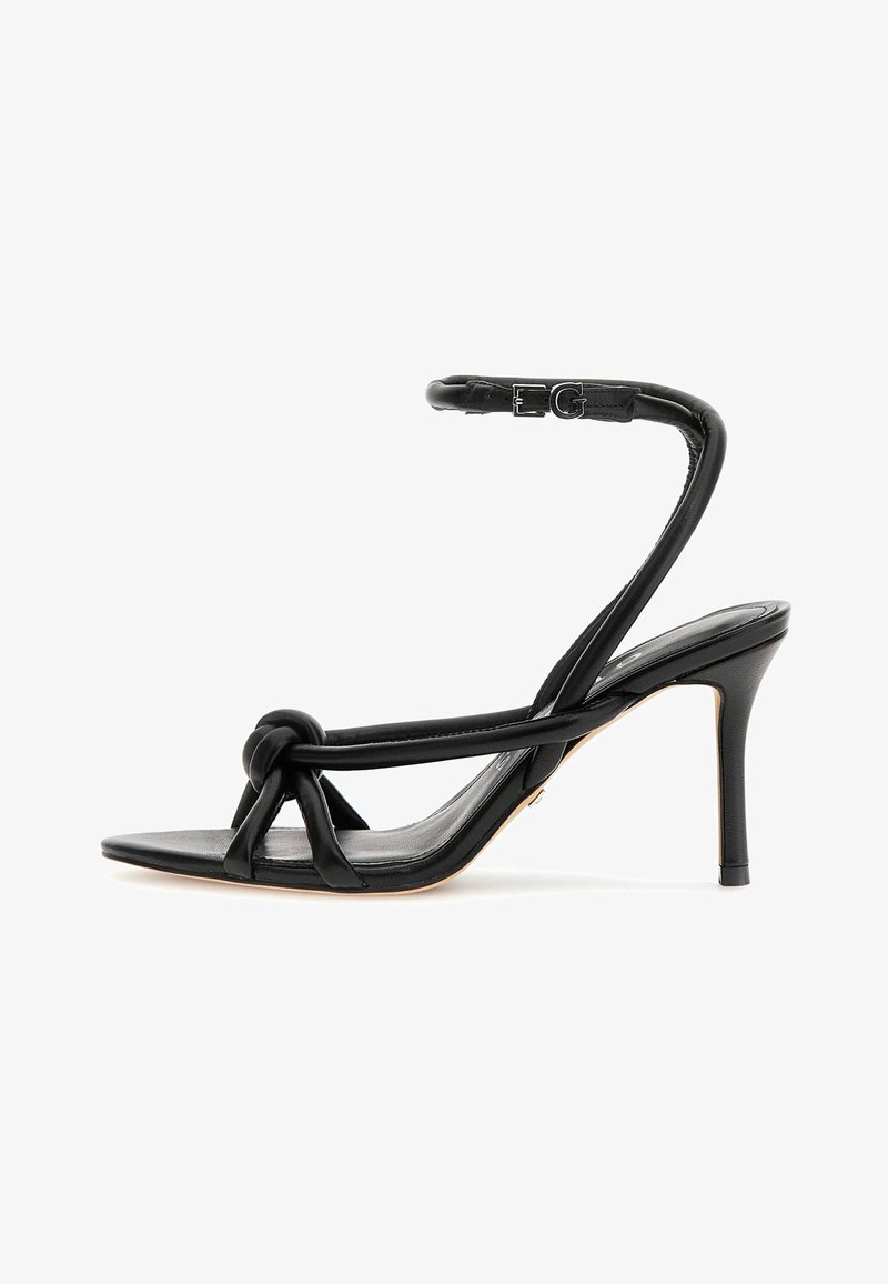 Sandalo con tacco alto in pelle nera, con un dettaglio a nodo davanti, cinturino alla caviglia con fibbia e design a punta. Texture liscia.