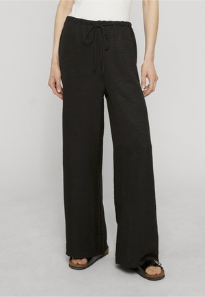Femme portant un pantalon noir ample à cordon et des sandales noires avec un haut blanc, debout devant un fond gris clair uni.