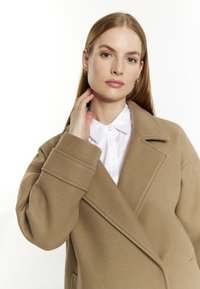 Beige wollen jas met een sjawlkragen, oversized pasvorm en omgeslagen manchetten, gelaagd over een wit overhemd met kraag. Glad textuur en minimalistisch ontwerp.