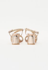 Talons dorés avec une sangle texturée, un talon en bloc métallique et une boucle à la cheville. Le design présente une finition lisse et brillante.