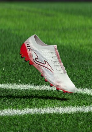 White and red soccer cleat with black studs hovering above green grass near white field boundary line.