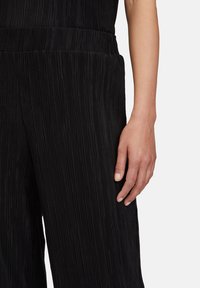 Černý plisovaný oděv s texturovanou tkaninou; pás je hladký. Ruka spočívá na boku a ukazuje přirozené nehty.