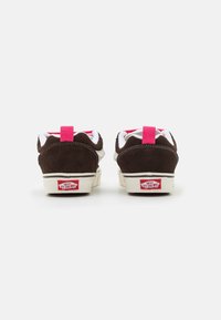 Ett par bruna Vans-sneakers med vita sulor, rosa hälflikar och röd "Vans Off The Wall"-logotyp på baksidan, visad från baksidan.