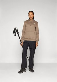 Felpa marrone con tasca frontale, pantaloni cargo neri e scarponi da trekking. Tiene in mano un bastone da trekking con impugnature grigie e cinghie nere.