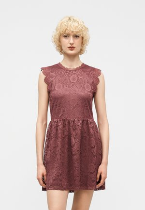 Giovane donna con capelli biondi corti e ricci che indossa un abito senza maniche in pizzo malva con motivi floreali, in piedi davanti a uno sfondo semplice.