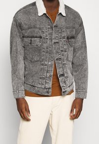 Giacca di denim grigia con colletto in pelliccia bianca, con due tasche sul petto e chiusura a bottoni. Indossata sopra una camicia marrone e pantaloni bianchi.