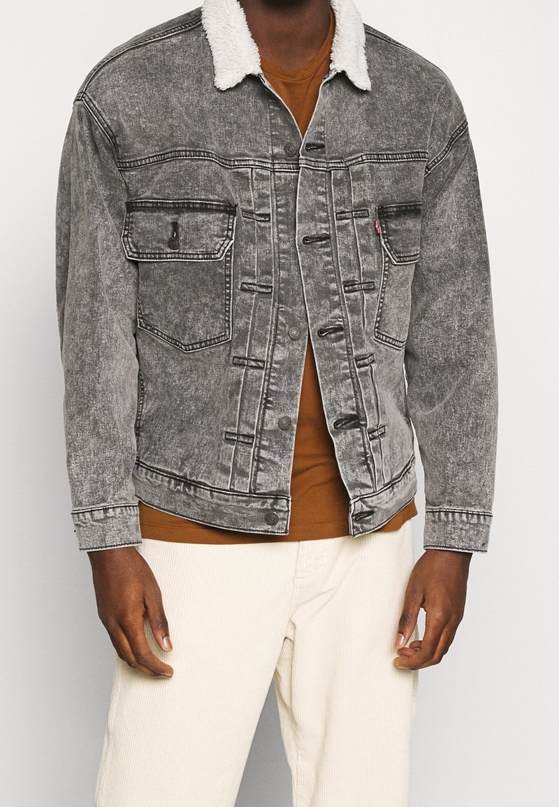 Giacca di denim grigia con colletto in pelliccia bianca, con due tasche sul petto e chiusura a bottoni. Indossata sopra una camicia marrone e pantaloni bianchi.