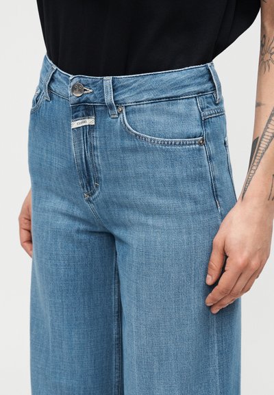 Άτομο που φοράει ανοιχτό μπλε τζιν ψηλόμεσο με μπροστινές τσέπες, μεταλλικό κουμπί και μικρή ετικέτα με την επωνυμία "Closed" ορατή στη μέση.