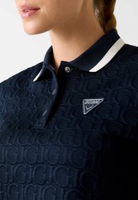 Granatowa koszulka polo z teksturowanym wzorem logo, białym kołnierzykiem w paski, zapięciem na zatrzaski oraz trójkątnym metalowym logo na piersi.