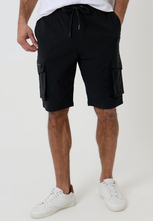 Mand iført sorte cargoshorts med snøre i taljen og hvide sneakers, står med hænderne i lommerne foran en ensfarvet baggrund.