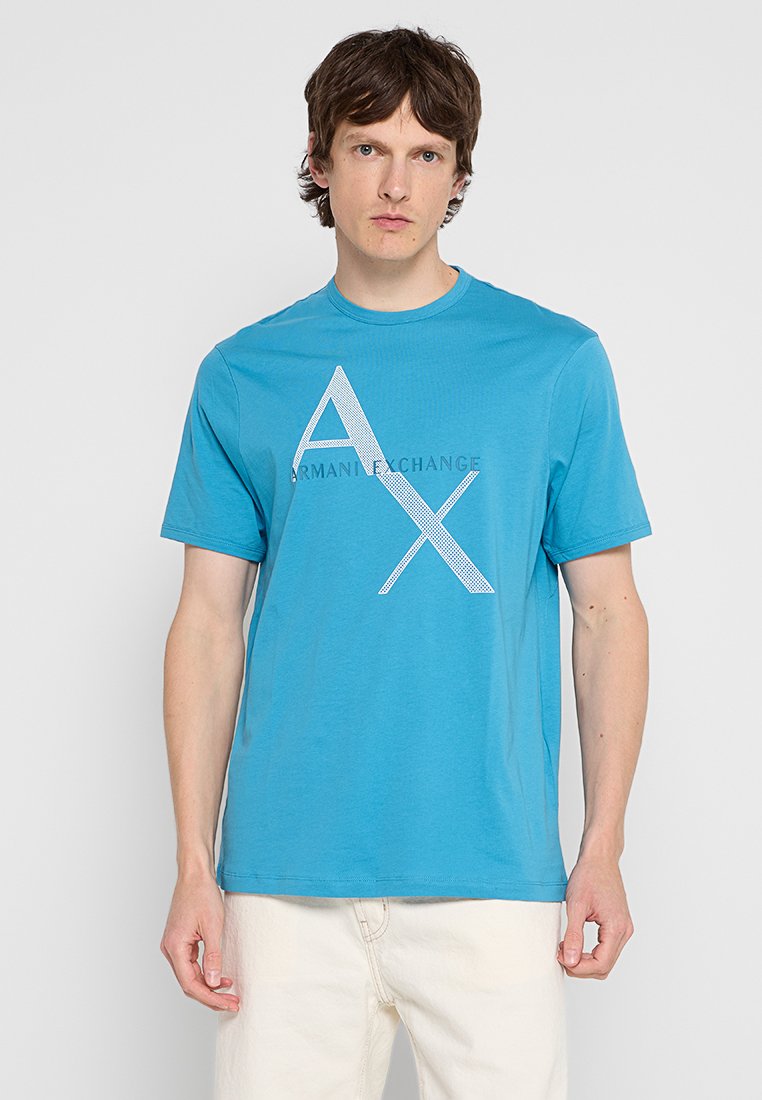 Uomo che indossa una maglietta turchese di Armani Exchange con il grande logo bianco "AX", abbinata a pantaloni beige chiaro, in piedi su uno sfondo semplice.