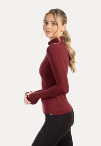 Long-sleeve fitted top in burgundy, featuring a high neck, stretchy fabric, and thumbholes at cuffs, worn over black leggings.