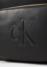 Geantă neagră din piele, având un logo „CK” proeminent și ridicat, cu un fermoar auriu, evidențiind o textură netedă și un design minimalist.