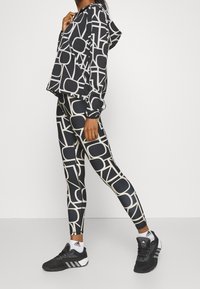 Gekürzte schwarze Kapuzenjacke mit cremefarbenen geometrischen Mustern, kombiniert mit passenden Leggings. Das Model trägt schwarze Sportschuhe.