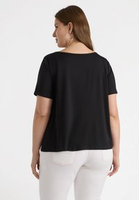 Top nero a maniche corte con una vestibilità rilassata, caratterizzato da un ampio scollo e una texture liscia. Abbinato a pantaloni chiari con cuciture a contrasto.