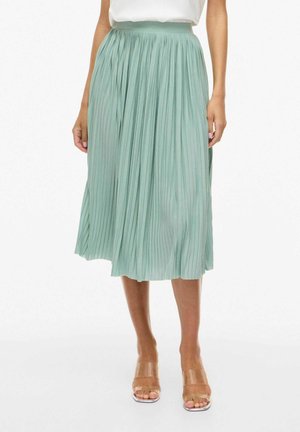 Femme portant une jupe midi plissée vert menthe, un haut blanc et des sandales transparentes à talons hauts, debout devant un fond blanc.