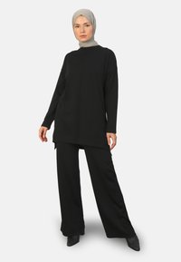 Zwarte lange mouwen tuniek met een hoge halslijn en zijsplitten, gecombineerd met zwarte wijde broek en zwarte puntige hakken. Glad textuur.