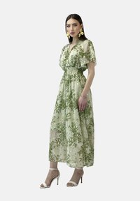 Maxikleid in Hellgrün mit einem Blumenmuster, geraffter Taille und kurzen, flatternden Ärmeln. Kombiniert mit weißen Absatzsandalen.