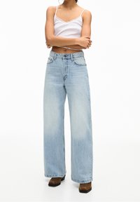 Personne portant un débardeur blanc avec dentelle, un jean taille haute large bleu clair, et des bottes marron à bout carré, debout les bras croisés.