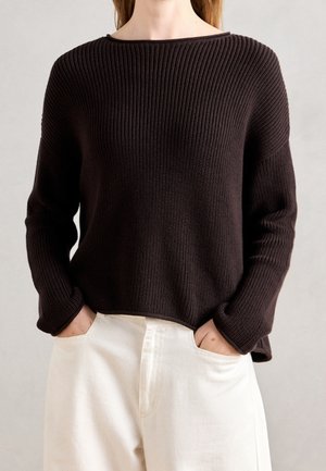 Personne portant un pull en tricot côtelé marron foncé à manches longues et un pantalon blanc, les mains dans les poches avant.