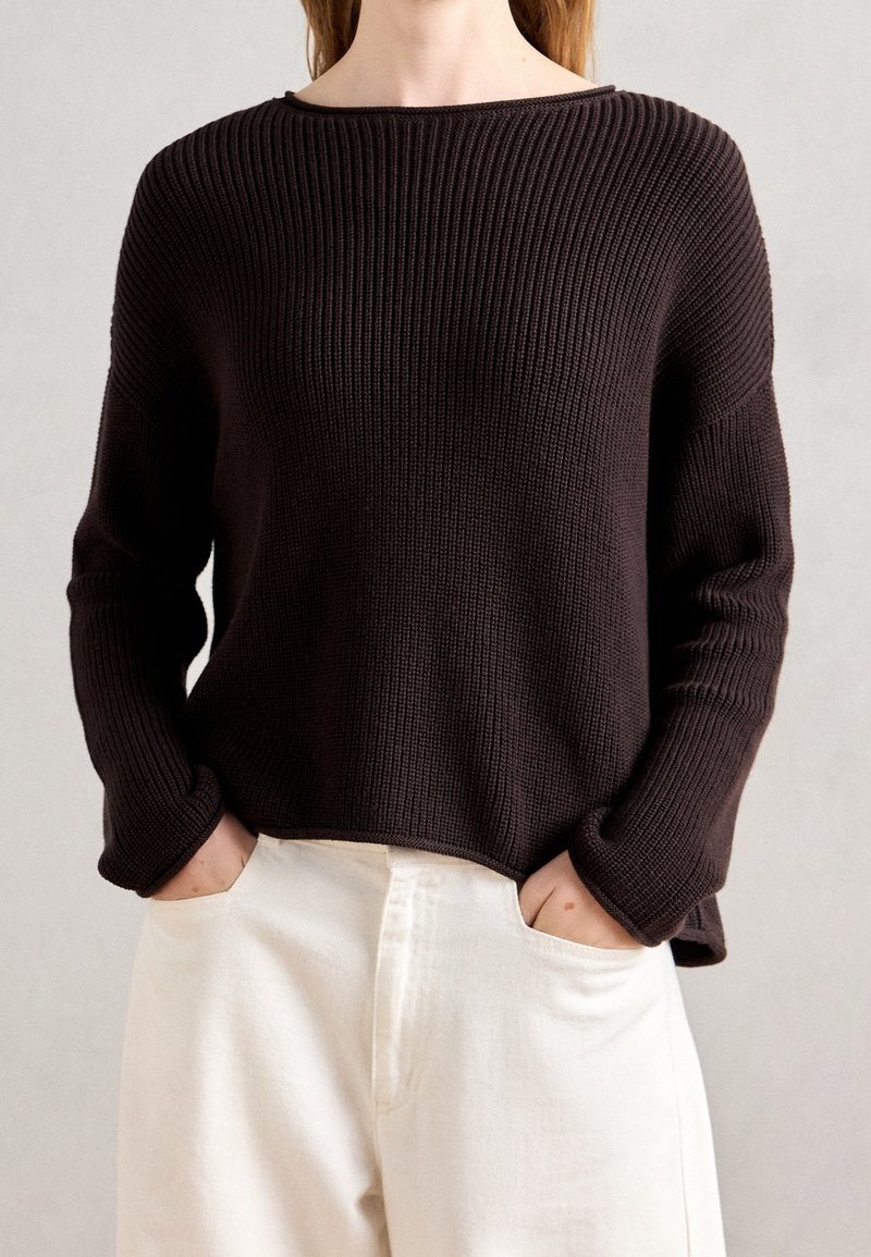 Personne portant un pull en tricot côtelé marron foncé à manches longues et un pantalon blanc, les mains dans les poches avant.