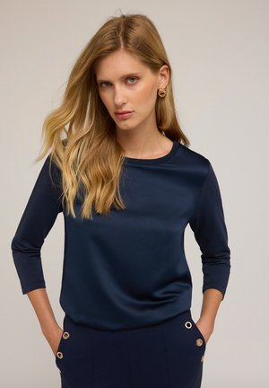 Frau mit langen blonden Haaren, die ein navyblaues Oberteil mit drei Viertelärmeln und passende Hosen mit goldenen Ösen trägt, mit Händen in den Taschen.