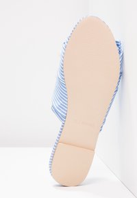 Semelle d'une chaussure plate rayée bleu et blanc avec une semelle beige lisse reposant contre un mur d'angle blanc.