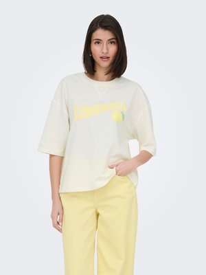 Femme portant un T-shirt crème oversize avec "LIMONATA" et un motif citron, associé à un pantalon jaune clair, debout sur un fond uni.
