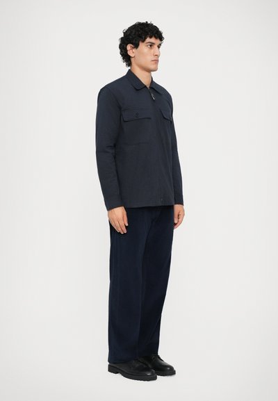 Navy blue jacket with two chest pockets, zip closure, and a collar, paired with matching wide-leg trousers and black shoes.