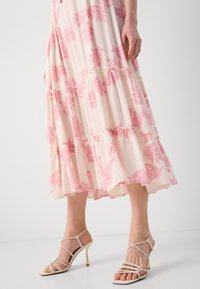 Bas des jambes portant des sandales beiges à talons hauts à brides et une robe blanche avec des motifs floraux feuillus roses et des volants superposés.