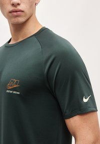 Άνδρας φοράει σκούρο πράσινο μπλουζάκι Nike με πορτοκαλί κείμενο "NIKE" και "Distant Dreams" στο στήθος, λευκό σήμα Nike στο μανίκι, κοντινό πλάνο.