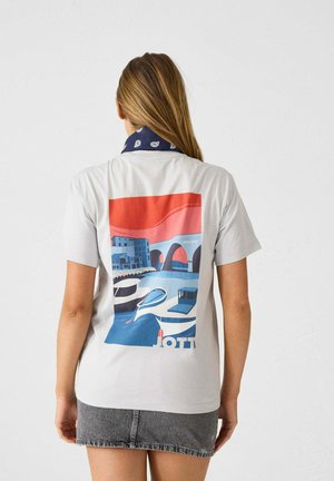 Camiseta gris claro con un diseño gráfico colorido en la parte posterior que representa una escena costera con edificios y barcos. Con mangas cortas.