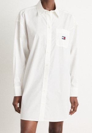 Modelka nosí dlouhé bílé košilové šaty s knoflíky a náprsní kapsou, na které je malé červené, bílé a modré obdélníkové logo.