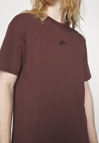 Bordový bavlněný tričko s krátkým rukávem a kulatým výstřihem. Na hrudi je vyšitá černá nášivka loga Nike. Jednoduchý, neformální design.