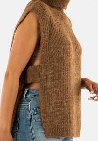 Pull en tricot sans manches marron avec des ouvertures latérales porté sur un jean bleu, montrant une bride de sous-vêtement beige sur un fond clair.