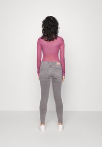 Vrouw met krullend zwart haar staat tegenover een witte muur, gekleed in een doorschijnende paarse lange mouwtop, grijze jeans en witte sneakers.