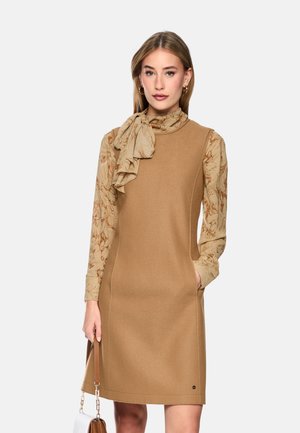 Young woman wearing a camel-colored sleeveless dress over a beige floral blouse with a neck bow, holding a white and brown handbag.