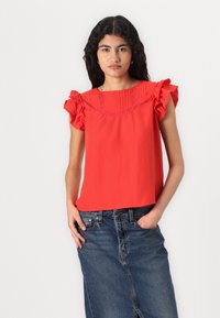 Rode top met gefronste mouwen, voorzien van een geplooide halslijn en geborduurde accenten. Gedirigeerd met een blauwe denim rok.