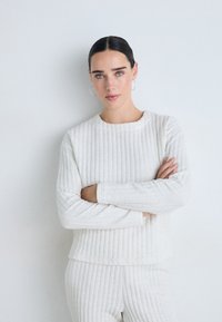Frau mit dunklen Haaren und blauen Augen trägt ein weißes, strukturiertes Langarmoberteil und eine Hose und steht mit verschränkten Armen vor einem schlichten weißen Hintergrund.