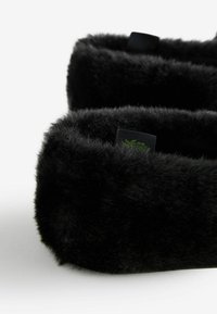 Czarna opaska na głowę z futra syntetycznego o miękkiej, puszystej fakturze. Posiada gładką wewnętrzną podszewkę oraz małą metkę z wzorem zielonego liścia.